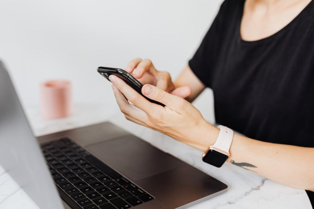 Naisoletetun kädet näppäilevät puhelinta. Taustalla pöydällä on kannettava tietokone ja kahvimuki.